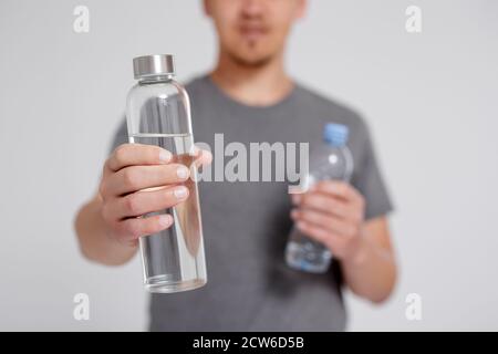 zero sprechi e concetto eco-friendly - giovane uomo a confronto acqua in vetro riutilizzabile e flacone di plastica su sfondo grigio Foto Stock