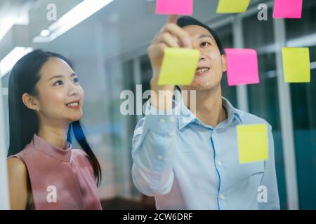 Due colleghi aziendali che attaccano post IT note su vetro, pianificazione, strategia, organizzazione Foto Stock