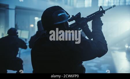 Ritratto di Masked Armed SWAT Police Officer Storming Dark sequestrato edificio di uffici con scrivanie e computer. I soldati con fucili e torce si muovono Foto Stock