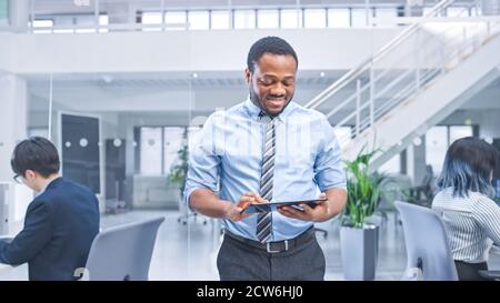 Handsome Young Black Manager in a Shirt Walking passa i suoi colleghi aziendali con un tablet e supervisiona il loro lavoro. Attività diversificate e motivate Foto Stock