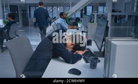 Young Japanese Manager è stanco al suo posto di lavoro con computer desktop e Falls addormentato. Diverse persone aziendali multietniche lavorano su computer in Foto Stock