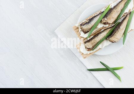 Sana colazione dietetica di pane di grano integrale con acciughe, formaggio cremoso, cipolla verde germogliata su sfondo di legno bianco, vista dall'alto, copia Foto Stock