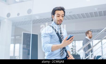 Giovane uomo d'affari felice entra in luminoso ufficio moderno, tiene smartphone e controlla i social media. In background diversi team di professionisti Foto Stock