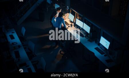 Tarda notte in ufficio moderno: Team diversificato di uomini d'affari di successo e donne d'affari si riuniscono intorno al computer, hanno discussione, trovare problema Foto Stock