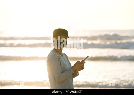 Ritratto di una donna che guarda la fotocamera con un cuffia nera e maschera facciale che chiacchiera con il suo telefono cellulare su un tramonto sulla spiaggia durante una vacanza in Foto Stock