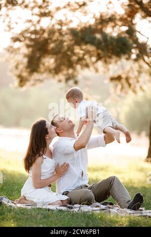 mamma, papà alza in alto il suo bambino a mezz'aria e la guarda sorridente. Felici genitori che trascorrono il tempo a giocare con il figlio nel parco il giorno d'estate. Selettivo Foto Stock