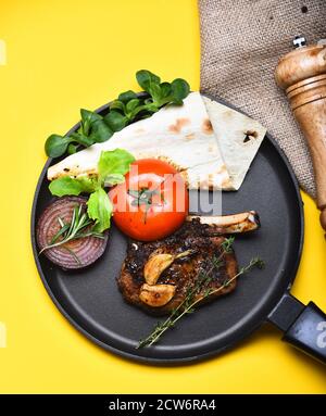 Costolette di maiale o agnello con decorazioni su padella, sfondo giallo. Bistecca arrosto con pomodoro, pane piatto e cipolla alla griglia. Concetto di cucina tradizionale. Piatti americani serviti nel ristorante o nella Steakhouse. Foto Stock