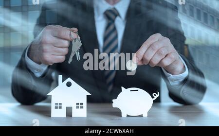 Uomo d'affari che tiene le chiavi e la moneta sopra una casa di carta e una banca del piggy della carta; esposizione multipla Foto Stock