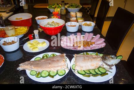 Un buffet di cibi freddi misti allestiti in una cucina preparata per una festa, tra cui un salmone intero con fettine di cetrioli, salumi e salsicce Foto Stock