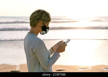 Ritratto di una donna in una maschera facciale che parla il suo telefono cellulare su una spiaggia al tramonto durante una vacanza in mezzo alla pandemia del coronavirus del cocid19 Foto Stock