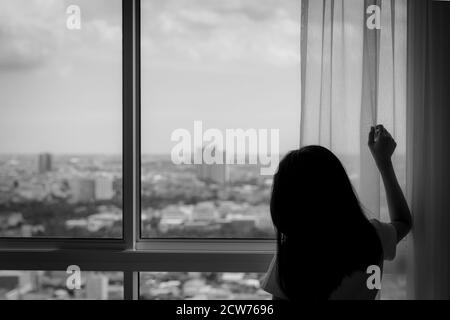 Vista posteriore della donna che tiene la tenda e che guarda fuori dal finestrino di vetro. Donna depressa da blocco e quarantena da pandemia di coronavirus. Mentale Foto Stock