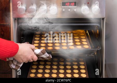 Mano che prende i biscotti caldi della noce fuori del forno a convezione fumante della stufa da cucina. Pasta frolla appena sfornata in stampi da forno in silicone su lamiera metallica. Foto Stock