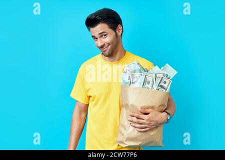 Divertente uomo tenere sacchetto di carta pieno di pile di denaro isolato su sfondo blu. Concetto di successo, di affari e di fortuna Foto Stock