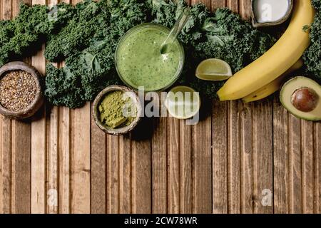 Bicchiere di frullato vegano verde sano, paglia di vetro. Ingredienti sopra. Kale, banane, avocado, calce, latte non-diario, polvere di matcha e semi sul legno Foto Stock