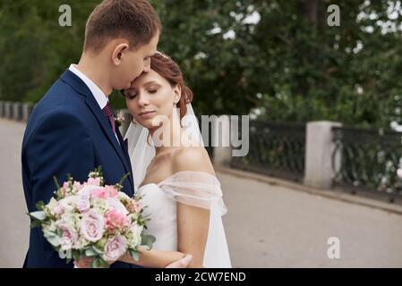 Un giovane bacia la sposa sulla fronte. Una giovane coppia felice si agguanata in un parco verde il giorno del loro matrimonio. Foto Stock