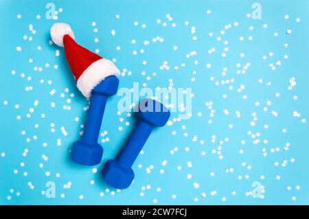 Piatto natalizio con manubri in cappello rosso di Babbo Natale su sfondo blu. Natale e Capodanno concetto di vacanza per fitness, allenamento e sano Foto Stock