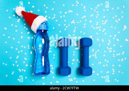 Piatto di Natale con manubri e bottiglia d'acqua nel cappello rosso di Babbo Natale su sfondo blu con fiocchi di neve. Natale e Capodanno concetto di vacanza Foto Stock