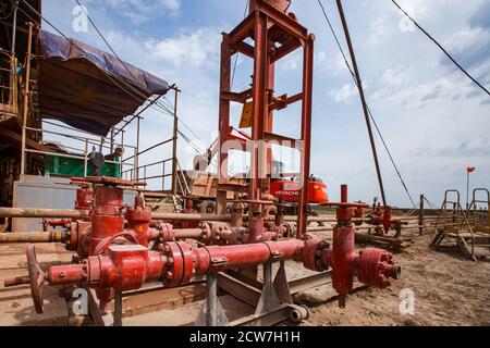 Deposito di olio Zhaikmunai nel deserto. Attrezzature per carri di perforazione. Primo piano delle valvole e dei tubi rossi. Escavatore Hitachi e dumper in background Foto Stock