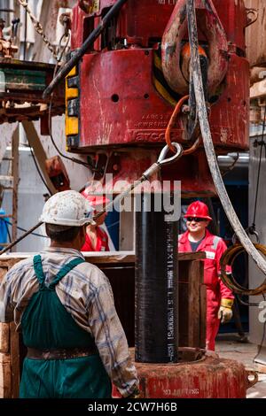 Operatore che utilizza un cavo in acciaio con gancio a scatto sul carro di perforazione. Motore elettrico e paranco. Lavoratori in usura rossa sfocati sullo sfondo. Deposito di olio di Zhaik-Munai Foto Stock