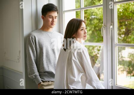 giovane coppia asiatica in piedi dalla finestra che parla chattando durante soggiorno a casa ordine Foto Stock