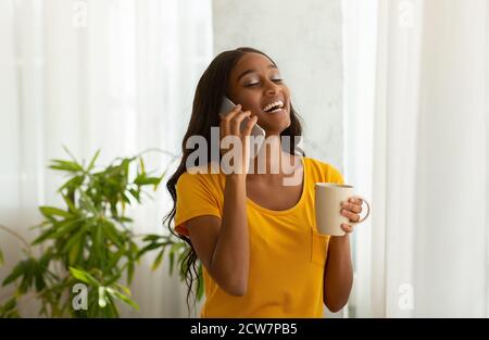 Donna nera di charme che parla sul cellulare con tazza di aromatico caffè a casa Foto Stock