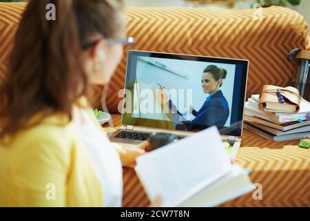 Visto da dietro donna in giacca gialla con cuffie bianche e libro di testo studio on-line su un computer portatile in casa in giornata di sole. Foto Stock