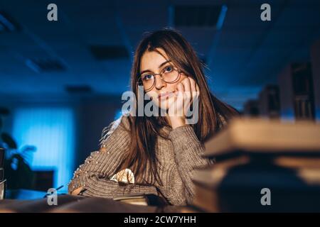 Stanca studentessa in serata seduta appoggiata sul braccio nella biblioteca alla scrivania con libri. Insegnamento e preparazione agli esami. Foto Stock