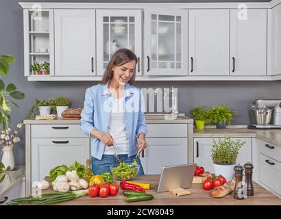 Giovane donna allegra che cucina in cucina con un tablet digitale Foto Stock