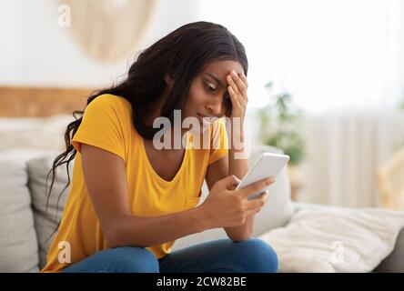 Infelice donna afroamericana guardando il suo smartphone con disgusto all'interno, spazio di copia Foto Stock