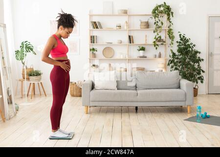 Infelice Afro-American Woman in piedi su bilance che toccano il ventriciolo Indoor Foto Stock