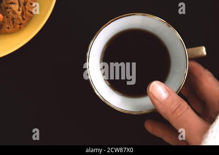 La mano della donna tiene una tazza di caffè in porcellana dorata su sfondo nero. Piatto giallo con biscotti aromatizzati con gocce colorate Foto Stock