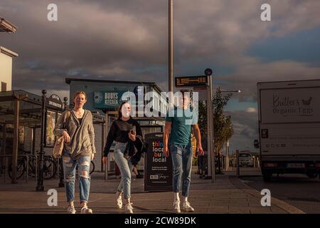 Vita quotidiana. Ragazze e uomini che camminano verso la macchina fotografica in via principale intorno alla stazione ferroviaria. Pietre di grigio. Irlanda. Foto Stock