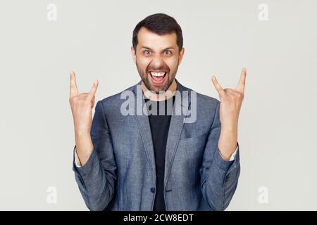 Giovane uomo d'affari con barba in giacca, gridando con un'espressione pazza sul viso, facendo un simbolo rock con le mani in su. Stella della musica. Concetto di musica pesante. Uomo su sfondo grigio. Foto Stock