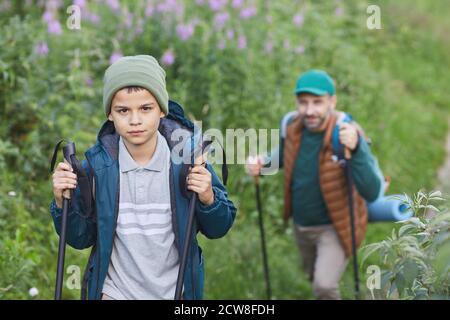 Ritratto di felice padre e figlio camminando insieme e camminando in salita, concentrarsi sul ragazzo sorridente con bastoni da trekking guardando la macchina fotografica in primo piano, copia spazio Foto Stock