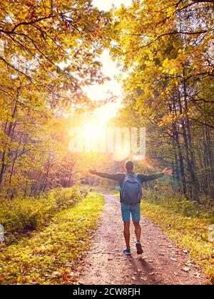Un ragazzo con braccia sollevate e uno zaino cammina lungo un sentiero nel parco autunnale contro una luce brillante nel corona di alberi Foto Stock