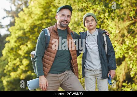 Ritratto di felice padre e figlio che camminano insieme e guardando la macchina fotografica mentre camminando nella foresta con zaino, copia spazio Foto Stock