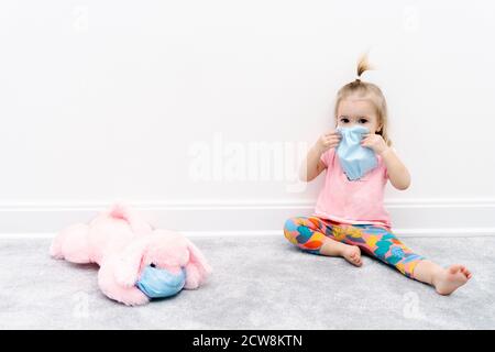 Piccolo bambino caucasico ragazza in maschera protettiva facciale si siede a. casa in quarantena con giocattolo rosa cane Foto Stock