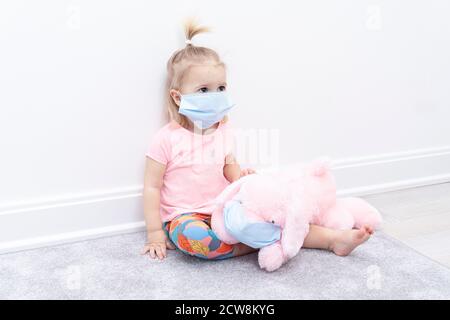Il bambino piccolo nella maschera protettiva facciale si siede a casa a. quarantena vicino al giocattolo rosa del cane Foto Stock