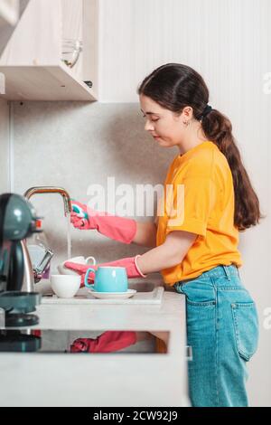 Lavori domestici. Una bella donna in abiti casual e guanti di gomma rosa è il lavaggio dei piatti. Vista laterale. Foto Stock