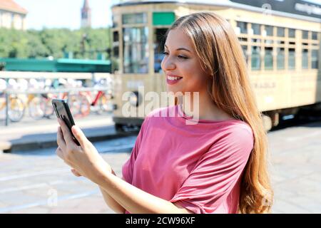 Bella giovane donna che tiene in mano lo smartphone in strada con il vecchio tram che passa sullo sfondo a Milano, Italia Foto Stock