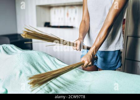 Massaggiatore femminile in t-shirt grigia e shorts blu che fanno massaggio con scopa da massaggio di bambù, cliente che è coperto con un grande asciugamano che giace sulla massa Foto Stock