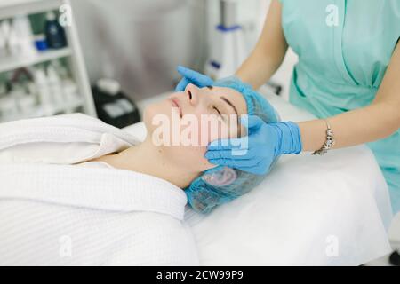 Cosmetoligist fare il massaggio del viso in salone di bellezza. Preparazione per l'iniezione. Cosmetologo in maschera medica protettiva Foto Stock