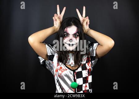 Donna in un costume halloween clown su sfondo nero isolato che posa divertente e pazzo con le dita sulla testa come orecchie conigliate, sorridendo allegro Foto Stock