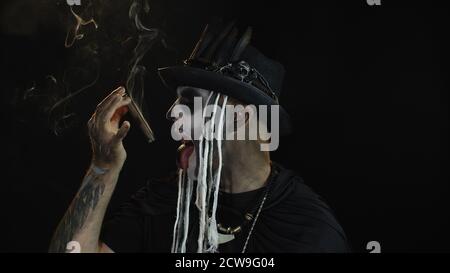 Ritratto di ragazzo spaventoso in costume di carnevale tematico di scheletro di Halloween contro sfondo nero fumando sigaro, facendo volti, mostrando lingua. Uomo sinistro con trucco professionale del cranio Foto Stock