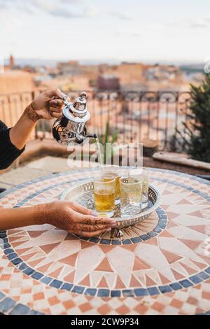 Mano donna che versa il tradizionale tè marocchino alla menta in bicchieri. Vassoio e teiera vintage color argento. Tavola rotonda a mosaico. Ospitalità del Marocco. Foto Stock