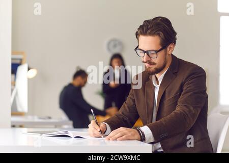 Giovane uomo d'affari seduto alla scrivania dell'ufficio che annotava gli obiettivi quotidiani o la strategia di lavoro nel diario di affari Foto Stock