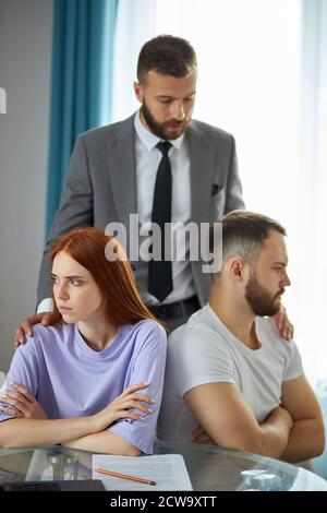 la donna rossa e l'uomo sopportato si siedono girandosi l'uno dall'altro, hanno litigato. psicologo fiducioso levarsi in piedi sopra la coppia, provi ad aiutare Foto Stock