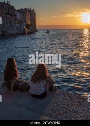 ROVIGNO, CROAZIA - 06/23/2018: Due giovani donne con capelli lunghi seduti vicino al mare e guardando il tramonto. Foto Stock