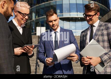 Giovane uomo d'affari che legge contratto con i suoi colleghi e che discutono si trova all'aperto vicino all'edificio dell'ufficio Foto Stock