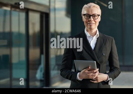 Ritratto di donna d'affari matura in occhiali e in nero elegante si adatta a tenere un tablet digitale e sorridere alla fotocamera all'aperto Foto Stock